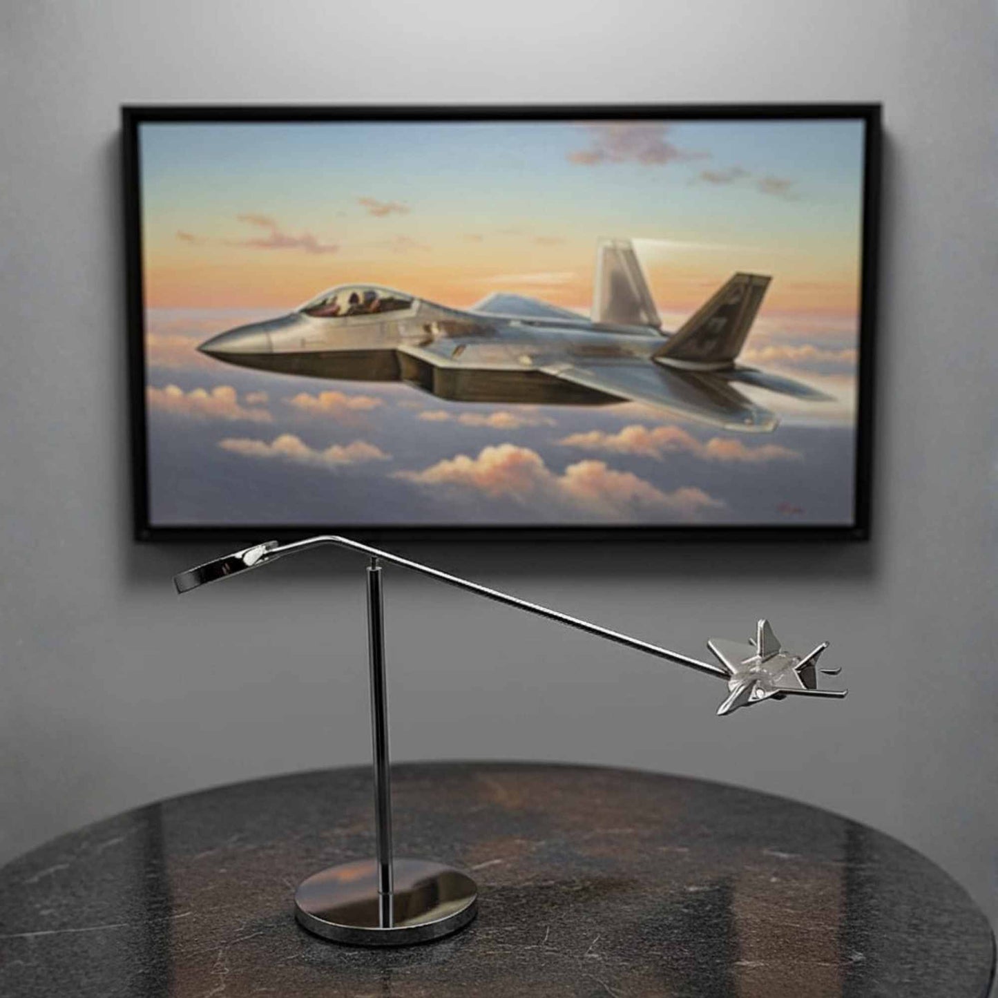 Framed artwork of a F22 Raptor fighter jet flying above clouds on a wall, with a metal stand model of a similar aircraft on a table.